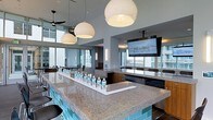 This contemporary amenity lounge is designed to impress, offering an open, airy layout bathed in natural light from expansive floor-to-ceiling windows that frame sweeping city skyline views. At the heart of the space sits a generously sized bar island topped with polished granite and accented by striking aqua tile along its base, providing ample seating for guests. Three large globe pendant lights hang overhead, casting a warm, inviting glow across the bar area and complementing the modern aesthetic. Mounted flat-screen televisions are positioned for easy viewing throughout the space, making it ideal for watching live events or presentations. A ceiling fan adds a casual, comfortable touch near the window-lined perimeter. The lounge transitions seamlessly into a separate dining or meeting area visible in the background, furnished with a long table and chairs, perfect for group gatherings or private events. Warm wood cabinetry and built-in appliances along the back wall add functional elegance. The overall ambiance is sleek and sophisticated, blending upscale design elements with a social, versatile atmosphere suited for both casual entertaining and more formal functions.