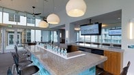 This contemporary amenity lounge is designed to impress, offering an open, airy layout bathed in natural light from expansive floor-to-ceiling windows that frame sweeping city skyline views. At the heart of the space sits a generously sized bar island topped with polished granite and accented by striking aqua tile along its base, providing ample seating for guests. Three large globe pendant lights hang overhead, casting a warm, inviting glow across the bar area and complementing the modern aesthetic. Mounted flat-screen televisions are positioned for easy viewing throughout the space, making it ideal for watching live events or presentations. A ceiling fan adds a casual, comfortable touch near the window-lined perimeter. The lounge transitions seamlessly into a separate dining or meeting area visible in the background, furnished with a long table and chairs, perfect for group gatherings or private events. Warm wood cabinetry and built-in appliances along the back wall add functional elegance. The overall ambiance is sleek and sophisticated, blending upscale design elements with a social, versatile atmosphere suited for both casual entertaining and more formal functions.