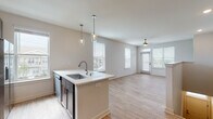 This well-appointed residence offers a thoughtfully designed open-concept layout that seamlessly connects the kitchen and living areas. The kitchen is a standout feature, showcasing rich dark cabinetry with sleek hardware, light quartz countertops, a white subway tile backsplash, and a full suite of stainless steel appliances. A central island with an undermount sink and stylish glass pendant lighting serves as both a functional workspace and a natural gathering point. Light wood-look flooring flows throughout the main living areas, enhancing the bright, airy atmosphere created by multiple large windows dressed with clean white blinds. The spacious living area is anchored by a ceiling fan and benefits from abundant natural light, with direct access to an outdoor space. A carpeted staircase with a clean white railing leads to the upper level, where a hallway landing with wood-look flooring connects to additional rooms. Bedrooms feature plush carpet, ceiling fans, and generous windows with neighborhood views. The bathroom is finished with wood-look tile flooring, dark cabinetry with a white countertop, and a glass-enclosed walk-in shower, offering a clean and functional design throughout.