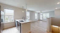 This well-appointed residence offers a thoughtfully designed open-concept layout that seamlessly connects the kitchen and living areas. The kitchen is a standout feature, showcasing rich dark cabinetry with sleek hardware, light quartz countertops, a white subway tile backsplash, and a full suite of stainless steel appliances. A central island with an undermount sink and stylish glass pendant lighting serves as both a functional workspace and a natural gathering point. Light wood-look flooring flows throughout the main living areas, enhancing the bright, airy atmosphere created by multiple large windows dressed with clean white blinds. The spacious living area is anchored by a ceiling fan and benefits from abundant natural light, with direct access to an outdoor space. A carpeted staircase with a clean white railing leads to the upper level, where a hallway landing with wood-look flooring connects to additional rooms. Bedrooms feature plush carpet, ceiling fans, and generous windows with neighborhood views. The bathroom is finished with wood-look tile flooring, dark cabinetry with a white countertop, and a glass-enclosed walk-in shower, offering a clean and functional design throughout.