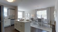 This single-floor residence showcases a thoughtfully designed open-concept layout that effortlessly connects the kitchen, dining, and living areas into one cohesive living space. The kitchen features a large central island with a crisp white quartz countertop, white shaker-style cabinetry with brushed nickel hardware, and dark hardwood flooring that anchors the space with warmth and contrast. The dining area is elegantly appointed with a glass-top table surrounded by upholstered chairs and illuminated by a classic chandelier pendant light, creating an inviting atmosphere for entertaining. A patterned area rug defines the dining zone, adding texture and sophistication. The living area transitions naturally from the dining space, highlighted by a plush white sofa and framed wall art that contribute to the refined aesthetic. Throughout the space, an abundance of natural light pours in through multiple large windows dressed with sheer, patterned curtains, enhancing the airy and luminous ambiance. The overall palette is a harmonious blend of whites and soft neutrals, giving the space a clean, modern, and welcoming character.