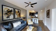 This well-appointed single-floor residence offers a thoughtfully designed open-concept layout that seamlessly connects the living area to the kitchen. The living space is anchored by rich hardwood-style flooring and features a ceiling fan with a sleek, modern fixture that adds both function and style. The walls are painted in a contemporary gray tone, complemented by bold abstract artwork that adds a sophisticated touch to the overall aesthetic.

The kitchen is a standout feature, showcasing dark espresso cabinetry, stainless steel appliances, and a peninsula with bar seating — ideal for casual dining or entertaining. Pendant lighting hangs above the counter, adding warmth and visual interest to the space.

The open layout creates an inviting flow, with the living area offering ample room for comfortable seating arrangements. Natural transitions between spaces are enhanced by consistent flooring and a cohesive neutral color palette accented with gold and navy tones. The entryway is visible from the main living area, providing a sense of depth and connectivity throughout the floor plan. Overall, the space balances modern design with a warm, livable ambiance.