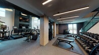 Located at 3560 Rambla Pl in Santa Clara, CA, this contemporary two-story fitness center is designed to deliver a premium workout experience across two well-appointed levels. The space is anchored by a dramatic double-height main floor, where soaring ceilings and expansive floor-to-ceiling windows flood the area with natural light and offer open views of the surrounding neighborhood. Patterned accent walls and linear LED lighting fixtures lend a sleek, modern aesthetic throughout.

The main level is equipped with a robust selection of cardio machines, including Matrix stair climbers and rowing machines, alongside a comprehensive free weights area featuring Hoist-branded dumbbells, adjustable benches, and a multi-station cable machine. Wood-accented columns and dark, textured rubber flooring add warmth and functionality to the space.

A glass-railed staircase with integrated step lighting connects the floors, leading to an upper level that houses a dedicated cycling studio with multiple Peloton-style spin bikes. Large mirrors on the upper floor enhance the sense of space, while the open mezzanine design allows views down to the main workout area below. A hydration station with a bottle-filling fountain is conveniently located near the stairwell. The facility is thoughtfully designed to support a full range of fitness activities in a stylish, well-lit environment.