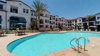 This residential unit is set within a multi-story complex showcasing Spanish Colonial-inspired architecture, characterized by white stucco exteriors, arched entryways, teal shutters, and terracotta-toned rooflines. The community's outdoor amenities are a standout feature, centered around a generously sized swimming pool with a smooth paver surround, lounge seating, and shade umbrellas, all framed by mature palm trees and abundant natural sunlight.

Inside, the bedroom serves as the focal point of the unit's interior. The room features neutral-toned carpet, soft white walls, and a ceiling fan, creating a calm and comfortable atmosphere. Natural light filters in through windows fitted with horizontal blinds. A tufted upholstered headboard with coordinating teal bedding adds a refined decorative touch, complemented by contemporary artwork displayed above the bed.

The bedroom includes two reach-in closets with built-in shelving, providing ample organized storage. A dedicated dresser area with a large framed mirror offers additional functionality. A hallway connects the bedroom to the rest of the unit, lined with recessed lighting and framed wall art. Decorative accents throughout, including ceramic vases and sculptural greenery, contribute to the cohesive, modern aesthetic of the space.