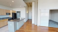 This single-floor loft-style space showcases an industrial-modern design defined by dramatically high ceilings with exposed concrete beams and visible ductwork, lending the unit a distinctive architectural character. The open-plan living area is anchored by warm hardwood flooring that flows seamlessly from the kitchen through to the main living space, creating a bright and cohesive environment.

The kitchen is well-appointed with light maple cabinetry, dark laminate countertops, a center island with a built-in cooktop, black appliances including a refrigerator and dishwasher, and tile flooring. An in-unit washer and dryer are conveniently located adjacent to the kitchen area.

Large, multi-pane windows throughout the space flood the interior with natural light and frame views of the surrounding urban landscape. The bedrooms feature plush carpet flooring and benefit from the soaring ceiling height, with exposed beam detailing adding to the loft aesthetic.

Two bathrooms serve the unit: one features a full soaking tub and shower combination with a warm-toned vanity countertop, while the second offers a stand-alone shower stall with a pedestal sink. Both bathrooms are finished with neutral tile flooring. Structural columns and a sleek sliding translucent panel add to the space's modern industrial sensibility throughout.