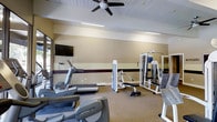 This fitness center at 7100 East Evans Avenue in Denver, Colorado offers a well-equipped, single-floor exercise space designed to support a variety of workout routines. The room features a selection of cardio machines, including treadmills and an elliptical trainer, alongside a multi-functional strength training machine and a flat workout bench, providing options for both cardiovascular and resistance training. A large wall-mounted flat-screen television is positioned for easy viewing during workouts, while a ceiling fan overhead helps maintain airflow and comfort throughout the space. Full-length mirrored panels line one wall, enhancing the sense of openness and allowing users to monitor their form during exercise. The flooring combines durable carpet and rubberized matting, offering appropriate surfaces for both machine-based and floor exercises. Neutral-toned walls with a distinctive accent stripe add a clean, structured aesthetic to the room. Natural light filters in from windows along one side, contributing to a bright and inviting atmosphere. The overall layout is thoughtfully organized to maximize usable floor space while keeping equipment accessible, making this fitness center a practical and functional amenity.