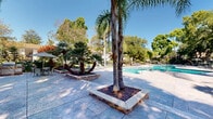 Located at 444 Saratoga Ave in Santa Clara, California, this outdoor amenity space offers a well-appointed, resort-style environment designed for relaxation and recreation. At its centerpiece is a generously sized swimming pool with a curved, freeform shape and clear turquoise water, surrounded by a broad, tiled pool deck ideal for lounging. Patio seating with shade umbrellas provides comfortable spots for outdoor dining and leisure.

The surrounding landscape is lush and thoughtfully designed, featuring a mix of mature shade trees and tall tropical palms that create a sense of privacy and seclusion. Raised brick-bordered planters showcase colorful ornamental plants and tropical foliage, adding vibrant contrast to the neutral stone paving. Large-format tile pavers extend throughout the outdoor area, creating a cohesive and polished surface that connects the pool zone to additional seating and relaxation areas.

Wrought iron fencing encloses the perimeter, providing security while maintaining an open, airy feel. The residential buildings visible in the background reflect a classic California-style architecture with warm stucco exteriors and covered balconies. Abundant natural sunlight and mature tree canopy together create a comfortable, shaded microclimate, making this outdoor space a welcoming retreat in the heart of Santa Clara's mild Bay Area climate.