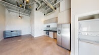 This single-floor urban loft delivers an industrial-modern aesthetic defined by its dramatic soaring ceilings with exposed steel beams and ductwork, complemented by sleek pendant lighting that adds a contemporary edge. Polished concrete floors run throughout the open-plan living area, reinforcing the loft's clean, minimalist character.

The well-appointed kitchen is a standout feature, outfitted with stainless steel appliances including a side-by-side refrigerator with water dispenser, electric range, built-in microwave, and dishwasher. Flat-panel cabinetry in a warm neutral tone extends to ceiling height, maximizing storage, while dark countertops and a subway tile backsplash complete the modern aesthetic.

A dedicated laundry nook houses a stacked washer and dryer unit, adding everyday convenience within the compact footprint. The two bathrooms are finished with crisp white cabinetry, concrete-toned countertops, hexagonal tile flooring, and a glass-enclosed walk-in shower with full white tile surround, offering a clean and functional design.

The open living area features smooth white walls, ample vertical space, and polished concrete underfoot, creating a versatile canvas suited to a variety of layouts. The overall ambiance is contemporary and urban, balancing industrial architectural details with refined finishes throughout.