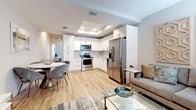 This well-appointed single-floor residence offers a thoughtfully designed layout that balances comfort and modern style. The open-concept living and dining area flows seamlessly into the kitchen, creating an inviting communal space ideal for both relaxing and entertaining. Light hardwood flooring runs throughout, complementing the soft, neutral color palette that defines the interior.

The kitchen is a standout feature, equipped with a full suite of stainless steel appliances — including a French door refrigerator, range, microwave, and dishwasher — set against crisp white shaker cabinetry and light countertops. A recessed tray ceiling with embedded lighting adds an elegant architectural detail overhead. The living area benefits from natural light through window treatments and is accented by distinctive geometric wall art panels.

The primary bedroom is a serene retreat, featuring a tray ceiling, built-in closet, and soft ambient lighting. A secondary bedroom offers similar comfort with its own closet and natural light. Both bathrooms are finished in clean white tones, each featuring a shower-tub combination, modern chrome fixtures, a single-sink vanity with ample storage, and a large mirror. A hallway connects the sleeping quarters to the main living space, maintaining a logical and practical flow throughout the residence.