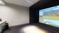 This golf simulator room is a purpose-built indoor space designed to deliver an immersive golfing experience regardless of weather or season. The room is finished with neutral grey walls and dark accent paneling that frames the large projection screen, creating a focused and distraction-free environment for players. The hitting area is positioned centrally, featuring a designated mat that serves as the tee and striking zone, illuminated by overhead LED strip lighting that ensures consistent visibility across the playing surface. The projection screen dominates one wall, displaying detailed course visuals — including fairway layouts, scorecard data, and environmental elements — providing a realistic simulation of real-world golf courses. The dark carpeted flooring absorbs impact and contributes to the room's controlled acoustic environment. The overall design is clean and functional, with a layout optimized for swing freedom and accurate shot tracking. This space is well-suited for individual practice sessions, lessons, or recreational play, offering a compact yet fully equipped setting for golfers of all skill levels to refine their game.