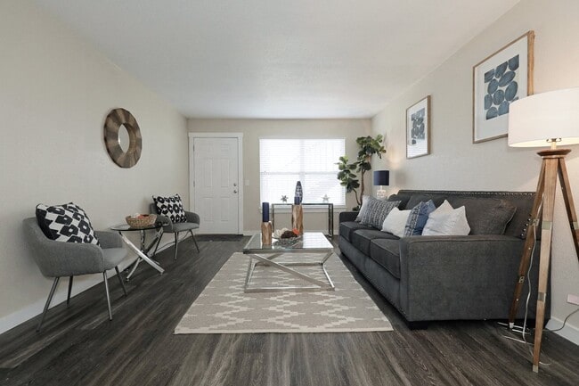Living room with dark hardwood floors, gray sofa and accent chair, glass coffee table, patterned rug, and framed artwork - Bennett Pointe