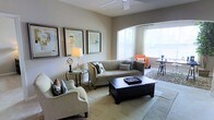 This single-floor space exudes a refined yet comfortable ambiance, thoughtfully designed to maximize both style and functionality. The living area serves as the centerpiece, featuring a neutral color palette of warm beiges and soft grays that creates a calm, welcoming atmosphere. A large arched window floods the room with abundant natural light, enhancing the airy, open feel of the space. The living room is furnished with a plush gray sofa, a classic cream armchair, and a bold dark-wood coffee table with built-in storage drawers, striking an elegant contrast against the light flooring. An accent armchair in a warm orange tone adds a pop of color near the window, complementing the sophisticated décor. The walls are adorned with a curated gallery of framed abstract artwork, adding a contemporary artistic touch. A ceiling fan overhead provides both practical comfort and a clean architectural detail. A round glass-top side table with a stylish table lamp rounds out the seating arrangement. The flooring transitions subtly across the space, with a textured area rug anchoring the main seating zone. Overall, the space balances modern design sensibilities with warm, livable comfort.