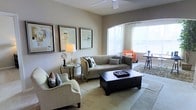 This single-floor space exudes a refined yet comfortable ambiance, thoughtfully designed to maximize both style and functionality. The living area serves as the centerpiece, featuring a neutral color palette of warm beiges and soft grays that creates a calm, welcoming atmosphere. A large arched window floods the room with abundant natural light, enhancing the airy, open feel of the space. The living room is furnished with a plush gray sofa, a classic cream armchair, and a bold dark-wood coffee table with built-in storage drawers, striking an elegant contrast against the light flooring. An accent armchair in a warm orange tone adds a pop of color near the window, complementing the sophisticated décor. The walls are adorned with a curated gallery of framed abstract artwork, adding a contemporary artistic touch. A ceiling fan overhead provides both practical comfort and a clean architectural detail. A round glass-top side table with a stylish table lamp rounds out the seating arrangement. The flooring transitions subtly across the space, with a textured area rug anchoring the main seating zone. Overall, the space balances modern design sensibilities with warm, livable comfort.