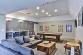 Located in Parkwood Circle SE in Atlanta, Georgia, this single-floor community game room is designed to offer residents a dynamic and engaging recreational experience. The space is thoughtfully divided into distinct activity zones, each contributing to a lively yet comfortable atmosphere.

At the heart of the room sits a full-size billiards table with a wood-tone frame, surrounded by ample space for gameplay. Colorful framed sports-themed artwork lines the walls, adding energy and visual interest to the area. Multiple wall-mounted flat-screen TVs are positioned throughout, ensuring excellent viewing from nearly every angle. A granite-topped bar counter with bar stools provides a casual spot to gather, complemented by low-profile upholstered bench seating nearby.

The lounge area features generously sized dark sectional sofas paired with a distinctive live-edge wood coffee table, creating a relaxed setting for conversation or watching TV. Accent pillows in warm amber tones add a welcoming pop of color. Natural light filters in through sliding glass doors, offering views of the surrounding outdoor area.

An adjacent arcade zone, complete with classic stand-up arcade cabinets and bold wall graphics, adds a playful, nostalgic touch to the space. Vaulted ceilings with recessed lighting throughout lend an open, airy feel, while neutral-toned carpet ties the entire room together cohesively.