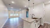 This single-floor unit offers a thoughtfully designed open-concept layout that seamlessly connects the kitchen and living area. The kitchen is a standout feature, showcasing a large center island with a quartz countertop, an undermount sink with a matte black faucet, and sleek pendant lighting overhead. White shaker-style cabinetry with black hardware lines the walls, complemented by a classic white subway tile backsplash and a full suite of stainless steel appliances, including a French door refrigerator, electric range, and over-the-range microwave. Wood-look luxury vinyl plank flooring flows consistently throughout the entire unit, adding warmth and continuity to the space. The open living area is generously proportioned and filled with natural light from a trio of large windows fitted with neutral roller shades, offering views of the surrounding landscape. Recessed lighting throughout ensures bright, even illumination in every room. The bathroom features a white vanity with quartz countertop, an undermount sink, and a framed mirror, with convenient access to a walk-in closet equipped with built-in shelving and hanging rods. The neutral color palette and modern finishes throughout create a clean, cohesive aesthetic well-suited for contemporary living.