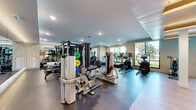 Located at 604 Vistera Blvd in Venice, Florida, this modern fitness center offers a thoughtfully designed, single-floor layout that accommodates a full range of workout options. The space is organized into distinct functional zones, allowing users to move seamlessly between cardio, strength training, and free weight areas.

The cardio section lines the perimeter near large windows, housing multiple treadmills, ellipticals, stair climbers, and stationary bikes — all positioned to take advantage of natural light and outdoor views of the surrounding Florida landscape. The strength training zone features a robust collection of cable machines, multi-functional cable systems, and weight benches, while full-length backlit mirrors along the walls visually expand the space and support proper form during workouts.

A dedicated free weights area includes neatly organized dumbbell racks alongside additional cardio equipment, mounted flat-screen televisions, and warm pendant lighting above a shiplap ceiling that adds a coastal design touch. Structural columns with clean white finishes divide this section while maintaining an open, airy feel.

Throughout the facility, recessed lighting ensures even illumination across polished concrete and light hardwood flooring. The contemporary, neutral color palette and generous glazing create a bright, motivating environment well-suited for a wide variety of fitness routines.