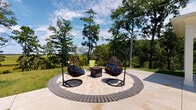 Located at 174 Overlook Point Pl in Charleston, SC, this single-level outdoor amenity space offers a thoughtfully designed retreat that blends natural surroundings with comfortable gathering areas. The centerpiece of the space is a circular paver patio, elegantly bordered by a contrasting brick ring and anchored by a central fire pit table. Two suspended wicker egg chairs with cushioned seating flank the fire pit, providing a stylish and relaxed setting ideal for socializing or quiet enjoyment. The patio is framed by mature trees and lush greenery, with expansive views stretching out to open marshland and the natural Lowcountry landscape beyond — a defining feature that sets this space apart. An adjacent concrete terrace extends the outdoor living area, furnished with additional wicker seating and offering a covered section with a full outdoor dining table and chairs, suitable for al fresco meals. A lawn area with Adirondack chairs and a cornhole game adds a recreational dimension to the space, while the neighboring multi-story residential building, finished in bold blue and white, provides a vibrant architectural backdrop. The combination of relaxed seating, natural views, and versatile gathering zones makes this outdoor space a well-rounded community amenity in the heart of coastal South Carolina.
