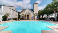 Located at 3952 Horseshoe Rd N in Little River, South Carolina, this single-story community clubhouse is a standout amenity within a well-maintained residential community. The building's exterior showcases a charming blend of beige vinyl siding and a stone-accented entryway, topped with a prominent peaked roofline and a tall chimney that add architectural character and visual height to the structure. Flanking the entrance, a pair of mature palm trees lend a coastal, subtropical feel that complements the South Carolina setting. The centerpiece of the outdoor space is a generously sized swimming pool with crystal-clear blue water, providing an inviting retreat for residents. A spacious pool deck surrounds the water, outfitted with lounge chairs and patio furniture, offering ample space for relaxation and socializing. A patio umbrella provides convenient shade for outdoor dining or lounging. The community is framed by lush, mature trees that provide natural privacy and greenery, enhancing the overall sense of seclusion and tranquility. With its clean, well-kept grounds and thoughtfully designed outdoor amenities, this space offers a resort-like atmosphere ideal for leisure and community gatherings.