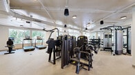 Located at 5875 Arapahoe Avenue in Boulder, Colorado, this well-equipped fitness center offers a generous, single-level layout designed to support a full range of workout routines. The space is thoughtfully organized to accommodate both strength training and cardiovascular exercise, with distinct zones flowing seamlessly throughout the open floor plan.

The strength training area is outfitted with a robust selection of professional-grade machines, including multi-station cable systems, a smith machine with free weights, and an extensive dumbbell rack covering a wide range of resistance levels. Adjustable benches are positioned throughout to complement the equipment. A large wall of mirrors enhances the sense of openness and allows users to monitor form during exercise.

The cardio zone features multiple treadmills with integrated digital displays, an elliptical trainer, a stair climber, and additional cardio equipment, all arranged near large windows that invite natural light and offer views of the surrounding tree-lined landscape. The bright, neutral-toned walls and resilient rubber flooring contribute to a clean and functional environment.

A dedicated open studio area with mirrored walls provides ample room for stretching, group classes, or bodyweight training. Exposed ductwork and pendant lighting fixtures lend an industrial-modern character to the space, while the overall layout ensures comfortable circulation throughout this comprehensive fitness facility.