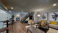 This well-appointed community amenity space combines contemporary design with functional areas, all unified by rich hardwood-style flooring and a textured ceiling with recessed lighting. The main lounge area serves as the heart of the space, furnished with mid-century modern seating — neutral upholstered chairs accented with bold yellow throw pillows — arranged around a central coffee table atop a plush patterned area rug. Large-scale abstract artwork adorns the walls, adding a vibrant artistic touch, while a striking arc floor lamp provides warm ambient lighting. A staircase with decorative tile risers and sleek black metal railings anchors the interior, creating a dramatic architectural focal point. Adjacent to the lounge, a dedicated business center offers dual computer workstations along a wall-mounted desk with bar-height seating, complemented by bold botanical artwork and a beverage station nearby. White modern chairs create an informal sitting area within this zone, bathed in natural light from a window. The space opens with an access door featuring frosted glass panels, enhancing the flow of natural light. Indoor plants are thoughtfully placed throughout, adding a fresh, inviting ambiance to this versatile and thoughtfully designed communal environment.