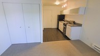 This single-floor unit presents an open-concept layout where the living area flows directly into the kitchen. The living space is finished with neutral wall-to-wall carpeting, lending a comfortable feel, while the kitchen features durable hardwood-style flooring that distinguishes the two zones. A set of full-height bifold closet doors along one wall provides generous storage capacity directly off the living area.

The kitchen is equipped with white cabinetry, a dark-toned countertop, a stainless steel gas range, and a black refrigerator. A single-basin sink is positioned against the back wall beneath the cabinetry, and overhead track lighting illuminates the space effectively. The layout allows for efficient use of the kitchen area without feeling cramped.

The unit's entry door is visible from the living space, contributing to the open and connected feel of the floor plan. Walls are finished in a clean white throughout, keeping the interior bright. Overall, the space offers a straightforward and functional design with practical finishes suited for everyday living.