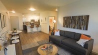 Located at 6750 E. Chenango Ave. in Denver, Colorado, this single-floor residence offers a thoughtfully designed open-concept layout that seamlessly connects the main living areas. The living room serves as a welcoming centerpiece, featuring warm hardwood-style flooring, a large sectional sofa, and a decorative wood-panel wall accent that adds texture and character to the space. A round wooden coffee table anchors the seating area, complemented by a striking floor lamp that provides ambient lighting.

The kitchen is a standout feature, showcasing sleek white cabinetry, stainless steel appliances, and a dark contrasting island with bar-height seating — ideal for casual dining or entertaining. Pendant lighting above the island adds a modern, polished touch, while the overall design prioritizes both function and style.

The open flow between the kitchen and living room creates a spacious, airy feel throughout the main area. A hallway visible from the living space leads to additional rooms, including what appears to be a bathroom with warm lighting. The neutral color palette, contemporary fixtures, and cohesive design elements come together to create a modern, comfortable, and highly functional living environment.
