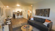 Located at 6750 E. Chenango Ave. in Denver, Colorado, this single-floor residence offers a thoughtfully designed open-concept layout that seamlessly connects the main living areas. The living room serves as a welcoming centerpiece, featuring warm hardwood-style flooring, a large sectional sofa, and a decorative wood-panel wall accent that adds texture and character to the space. A round wooden coffee table anchors the seating area, complemented by a striking floor lamp that provides ambient lighting.

The kitchen is a standout feature, showcasing sleek white cabinetry, stainless steel appliances, and a dark contrasting island with bar-height seating — ideal for casual dining or entertaining. Pendant lighting above the island adds a modern, polished touch, while the overall design prioritizes both function and style.

The open flow between the kitchen and living room creates a spacious, airy feel throughout the main area. A hallway visible from the living space leads to additional rooms, including what appears to be a bathroom with warm lighting. The neutral color palette, contemporary fixtures, and cohesive design elements come together to create a modern, comfortable, and highly functional living environment.