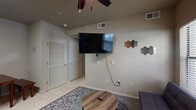 This single-floor unit presents a functional and well-organized layout across several distinct living spaces. The living area serves as the central hub of the unit, featuring light-toned tile flooring, a ceiling fan, and a wall-mounted flat-screen television. Hexagonal decorative wall panels add a modern touch, while a patterned area rug and wooden coffee table complete the casual, comfortable setting. An adjacent dining area provides space for meals with a compact wooden table and seating. The kitchen is efficiently designed with warm wood cabinetry, dark countertops, and a full suite of stainless steel appliances, including a refrigerator, electric range, and over-the-range microwave — offering everything needed for convenient meal preparation. The unit includes two bedrooms: one larger room with hardwood-style flooring, clean neutral walls, and a full-size bed with a dark-framed headboard, and a second, cozier bedroom featuring a twin bed, ceiling fan, and a large window fitted with horizontal blinds that allow natural light to filter in. The bathroom is finished with tile flooring, a toilet, and wood-toned upper cabinetry providing storage. Throughout the unit, neutral wall tones and consistent finishes create a cohesive, understated aesthetic that is both practical and inviting.