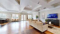 This generously proportioned single-floor community space makes a strong architectural impression through its distinctive coffered ceiling design, which adds depth and sophistication to the open layout. Richly stained concrete floors in warm amber and deep brown tones flow throughout, creating a striking visual contrast against the crisp white walls and trim.

The main lounge area is thoughtfully arranged with plush, neutral-toned sectional seating centered around a large flat-screen television, flanked by decorative artwork and lush indoor plants that bring a refreshing organic element to the space. Oversized French doors and large windows line the entry-facing wall, flooding the interior with natural light and offering views to the outdoors.

A dedicated seating nook near the entrance features accent chairs and a distinctive wicker hanging chair, creating a casual, welcoming reception area. Mirrored wall panels in this zone visually expand the already generous footprint.

A separate business center alcove is equipped with multiple computer workstations, modern pendant lighting, and additional window views, making it well-suited for productive work sessions. The overall ambiance blends contemporary design with warm, inviting elements — a polished yet comfortable environment suitable for both social gatherings and individual use.