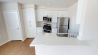 This single-story space showcases a well-appointed kitchen designed with a clean, contemporary aesthetic. The kitchen is outfitted with bright white shaker-style cabinetry that extends to the upper walls, maximizing storage capacity through a combination of upper cabinets, lower cabinets, and a tall pantry-style cabinet column. The cabinetry is complemented by sleek chrome hardware, contributing to the space's modern sensibility.

A suite of stainless steel appliances anchors the kitchen, including a gas range with oven, an over-the-range microwave, and a full-size refrigerator. The appliances are thoughtfully positioned within the cabinetry layout, creating an efficient and functional cooking zone. Light-toned quartz or quartz-style countertops provide generous workspace throughout, maintaining a cohesive, luminous palette.

A peninsula extends into the open area, featuring an undermount stainless steel sink with a contemporary chrome faucet. This peninsula serves as both a functional prep area and a natural divider between the kitchen and the adjoining living space. Light wood-tone flooring runs throughout, adding warmth against the predominantly white interior. The overall ambiance is clean, bright, and modern.