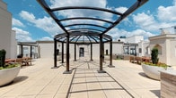 Located at 5555 E Mockingbird Ln in Dallas, Texas, this rooftop terrace offers an expansive open-air retreat with a thoughtfully designed layout ideal for gatherings and leisure. The centerpiece of the space is a dramatic circular pergola structure featuring a curved metal frame with a translucent glass canopy, allowing abundant natural light to filter through while providing partial shelter. Flanking columns lend an architectural elegance to the structure, casting geometric shadows across the large-format, light-toned stone tile flooring that extends throughout the terrace.

The outdoor kitchen area is appointed with a mosaic tile-clad counter, granite-style countertop surface, and an integrated grill, making it well-suited for outdoor entertaining. Nearby, a dedicated lounge zone features wicker seating alongside a built-in fireplace element framed by a stone surround, adding warmth and ambiance to the space. A wooden dining table with chairs provides an additional gathering area.

Decorative planters with manicured topiaries and seasonal flowers add greenery and character to the rooftop setting. The terrace is accessed via a metal utility door and enjoys open sky views, with the Dallas skyline and surrounding urban landscape visible in the background, enhancing the elevated atmosphere of this well-appointed outdoor space.