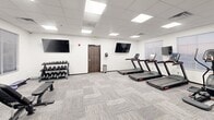Located at 1908 Fox Rd in Rapid City, South Dakota, this fitness center offers a thoughtfully organized and fully equipped gym space across a single floor. The room features a clean, modern aesthetic with white walls, a drop-panel ceiling with recessed flat-panel lighting, and durable patterned carpet tiles throughout.

Cardio equipment is well-represented, with multiple treadmills and elliptical trainers positioned along the walls, each accompanied by wall-mounted flat-screen TVs for entertainment during workouts. A stationary bike adds to the cardiovascular options available.

The strength training area includes a multi-functional cable machine, a weight bench, and a well-stocked dumbbell rack with a full range of free weights. A medicine ball and accessory rack rounds out the functional fitness offerings, providing versatility for a range of exercise routines.

Natural light filters into the space through multiple windows fitted with horizontal blinds, creating a bright and inviting atmosphere. The layout is open and efficient, allowing comfortable movement between equipment zones. A centrally positioned wall-mounted TV ensures visibility from multiple areas of the room. The overall design prioritizes functionality and cleanliness, making this gym a practical and accessible fitness facility.
