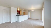 This single-floor space offers an open-concept layout that seamlessly connects the kitchen and living area, creating a cohesive and functional environment. The kitchen is outfitted with rich, warm-toned wood cabinetry and dark granite-style countertops that provide a striking contrast against the bright white walls. A peninsula-style breakfast bar extends into the living space, offering additional workspace and a natural dividing point between the two areas. Stainless steel appliances, including a microwave, are integrated into the kitchen design. The living area is finished with plush, neutral-toned carpeting that adds warmth and comfort underfoot. Recessed lighting in the kitchen is complemented by a stylish pendant light fixture over the living area, ensuring consistent, well-distributed illumination throughout. The walls are painted in a clean, crisp white tone, contributing to a bright and airy atmosphere. A doorway visible in the background suggests access to additional rooms or hallways, hinting at further functional spaces beyond the main open area. Overall, the layout balances practicality with a clean, modern aesthetic.