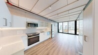 Located in the heart of Seattle's Belltown neighborhood, this unit at 3zero3 offers a thoughtfully designed open-plan layout that combines the living room and kitchen into one cohesive, functional space. Light wood-tone flooring runs throughout, complementing the clean, contemporary aesthetic.

The kitchen is well-appointed with white flat-panel cabinetry, open shelving, white quartz countertops, a tile backsplash, and stainless steel appliances including a GE range, over-the-range microwave, refrigerator, and dishwasher. Under-cabinet lighting adds a refined touch to the workspace.

The living area is defined by its striking floor-to-ceiling sliding glass doors, which open to a balcony and frame sweeping views of the Seattle skyline. Natural light floods the interior, enhancing the bright, airy atmosphere. A structured ceiling with an exposed grid detail and integrated track lighting adds an industrial-modern character to the space.

Along one wall of the living area, a built-in panel system with integrated storage and a wall-mounted HVAC unit maintains the streamlined design. The unit also features a smart lock entry. Compact yet efficient, this space maximizes every square foot with modern finishes and urban views that define Seattle living.