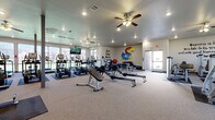 Located at 5401 Rock Chalk Dr in Lawrence, Kansas, this well-equipped fitness center spans a single floor with generous, open square footage designed to accommodate a comprehensive workout experience. The layout is thoughtfully organized, with dedicated zones for cardio, strength training, and functional fitness. A full row of True-brand treadmills and elliptical machines lines the perimeter near floor-to-ceiling windows, allowing users to exercise with an abundance of natural light and outdoor views of the surrounding community. Rowing machines and weight benches occupy the central floor space, providing ample room for functional training. A dumbbell rack, stability balls, and weight benches make up a focused free weights area, while additional selectorized strength machines round out the equipment offerings. Ceiling fans and recessed lighting maintain a comfortable and well-lit environment throughout. The space is unified by a strong University of Kansas Jayhawks theme, with a large KU Jayhawk mascot mural as a bold focal point, complemented by motivational wall art including phrases like "Never Give Up," "Believe Repeat," and "Fitness is a Lifestyle." Mounted flat-screen televisions add to the user experience. The carpeted flooring and rubber equipment mats contribute to a clean, functional aesthetic throughout this spirited community fitness facility.