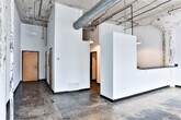 Located at 828 Ralph McGill Blvd NE in Atlanta's Old Fourth Ward neighborhood, this single-floor space embodies a raw industrial aesthetic that blends original architectural character with functional modern amenities. The open main area commands attention with its expansive concrete floors bearing the natural patina of age, exposed overhead ductwork and piping, and weathered structural columns that speak to the building's industrial heritage. Soaring ceilings amplify the sense of volume throughout, while large steel-framed factory windows allow abundant natural light to pour in and offer views of the surrounding streetscape. A kitchenette area features dark countertops, a stainless steel sink, white cabinetry, and a dishwasher — all set beneath pendant lighting suspended from the raw ceiling above. A dedicated vanity room includes a wide countertop with an integrated sink and a full-width mirror with overhead lighting, well-suited for use as a dressing or makeup area. The space also includes a separate laundry room equipped with a full-size washer and dryer. Throughout, the contrast of clean white walls against the aged concrete floors and distressed ceilings creates a distinctive, loft-like atmosphere ideal for a variety of creative or commercial uses.