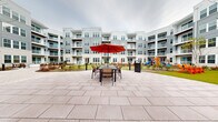 This expansive outdoor courtyard is the centerpiece of a multi-story residential community, designed to offer a diverse range of amenities within a beautifully landscaped environment. The courtyard is enclosed on all sides by the curved facade of the building, which features neutral gray and beige siding, private balconies with white railings, and warm wood-accented overhangs that lend architectural character to the exterior.

The paved courtyard area is composed of large-format stone pavers arranged in sweeping, curved pathways that guide residents through the various zones. Multiple seating areas are positioned throughout, each outfitted with tables, chairs, and bold red market umbrellas, creating inviting spots for outdoor dining and socializing. Vibrant orange Adirondack chairs are arranged on a well-maintained artificial lawn, providing a colorful and relaxed lounge area.

A putting green with a regulation flag offers a recreational focal point, surrounded by thoughtfully curated landscaping featuring ornamental shrubs and seasonal plantings in mulched beds. A children's play area complete with a colorful slide and swing set is nestled within the courtyard, making the space family-friendly. Pathway lighting via sleek bollard fixtures ensures the space is functional and welcoming at all hours.
