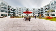 This expansive outdoor courtyard is the centerpiece of a multi-story residential community, designed to offer a diverse range of amenities within a beautifully landscaped environment. The courtyard is enclosed on all sides by the curved facade of the building, which features neutral gray and beige siding, private balconies with white railings, and warm wood-accented overhangs that lend architectural character to the exterior.

The paved courtyard area is composed of large-format stone pavers arranged in sweeping, curved pathways that guide residents through the various zones. Multiple seating areas are positioned throughout, each outfitted with tables, chairs, and bold red market umbrellas, creating inviting spots for outdoor dining and socializing. Vibrant orange Adirondack chairs are arranged on a well-maintained artificial lawn, providing a colorful and relaxed lounge area.

A putting green with a regulation flag offers a recreational focal point, surrounded by thoughtfully curated landscaping featuring ornamental shrubs and seasonal plantings in mulched beds. A children's play area complete with a colorful slide and swing set is nestled within the courtyard, making the space family-friendly. Pathway lighting via sleek bollard fixtures ensures the space is functional and welcoming at all hours.