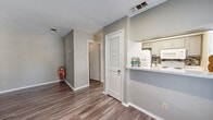 This single-floor unit offers an efficient, open-concept layout that combines the kitchen and living area into one cohesive space. The interior is finished with durable wood-look plank flooring throughout, providing a warm, consistent aesthetic across all areas. Neutral gray walls and crisp white trim create a clean, modern backdrop that enhances the sense of openness.

The kitchen is positioned along one wall and features a raised breakfast bar with a light granite-style countertop that opens toward the living area, promoting an easy flow between the two spaces. Gray cabinetry, a tile mosaic backsplash, white appliances including a refrigerator and microwave, and a single-basin sink with a modern faucet complete the kitchen's functional design.

The living area is well-lit by multiple windows fitted with white horizontal blinds, allowing ample natural light to fill the space. A louvered interior door adds a subtle architectural detail, while recessed lighting overhead provides additional illumination. The textured ceiling and multiple HVAC vents reflect practical construction. The overall ambiance is clean, understated, and move-in ready.