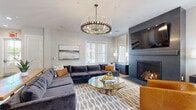 This thoughtfully designed single-floor space offers a seamless open-concept layout that connects the kitchen, dining, and living areas into one cohesive, flowing environment. Light hardwood flooring runs throughout, providing a warm foundation that complements the contemporary aesthetic.

The kitchen is a standout feature, anchored by an expansive island with a crisp white quartz countertop and a bold slate-blue base, surrounded by houndstooth-upholstered bar stools. White shaker-style cabinetry lines the walls, accented by a herringbone tile backsplash and a stainless steel range hood. A suite of stainless steel appliances and elegant pendant lighting complete the space.

The living area is generously proportioned and centers around a dramatic dark-surround fireplace with a wall-mounted television above. A large sectional sofa in deep charcoal velvet anchors the room, complemented by a cognac leather accent chair and a sculptural glass coffee table. A statement ring chandelier with exposed bulbs hangs overhead, adding a refined yet bold focal point.

Natural light filters in through multiple windows dressed with white blinds, creating a bright and airy atmosphere. French doors offer a view to the outdoors, further enhancing the sense of openness. The overall ambiance is modern and sophisticated, balancing comfort with carefully curated design details throughout.