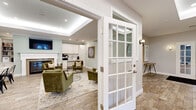 Located at 100 Town Walk Dr in Hamden, Connecticut, this single-story amenity center offers a comprehensive collection of community spaces designed to support an active and social lifestyle. The welcoming lounge serves as a central gathering area, featuring a tray ceiling, a classic fireplace with a white surround, a wall-mounted television, built-in bookshelves, and plush green velvet seating arranged around a glass coffee table. An adjacent kitchenette with white cabinetry provides convenient refreshment options, while a dining table offers additional seating. The expansive fitness center is well-equipped with treadmills, cable machines, a punching bag, and a dedicated row of elliptical trainers. A separate studio space features hardwood flooring, floor-to-ceiling mirrors, a ballet barre, and a Peloton bike, ideal for cycling or barre workouts. A stylish co-working lounge offers a long wooden desk along a blue accent wall, globe pendant lighting, and contemporary artwork. The children's area is thoughtfully designed with soft blue walls, colorful patterned carpeting, built-in shelving, play kitchens, bean bag seating, and natural light from multiple windows. Rounding out the amenities is a full-size indoor basketball court with high ceilings and perimeter windows, completing this versatile community facility.