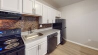 Located at 300 Audubon Pkwy in Syracuse, New York, this single-floor unit offers a thoughtfully updated interior with a mix of modern finishes and functional design. The kitchen is equipped with crisp white shaker-style cabinetry, a textured stone mosaic tile backsplash, light gray countertops, a stainless steel sink, and black appliances including a refrigerator and dishwasher. Light wood-look flooring runs through the kitchen and main living areas, contributing to a clean, contemporary feel. The living area is generously sized, featuring recessed ceiling lighting, a wall-mounted air conditioning unit, and a full wall of vertical blinds that allow for adjustable natural light. Subtle LED accent lighting along the base of the blinds adds a distinctive modern touch. The bedrooms feature plush carpeted flooring and neutral-toned walls, creating a calm and comfortable atmosphere. One bedroom benefits from a window with exterior views. The bathroom is well-appointed with marble-look tile flooring, a dual-toned tiled tub and shower surround combining white marble and dark gray stone-effect tiles, matte black fixtures, and a warm wood-toned vanity with a white countertop. Overall, the unit presents a cohesive, updated aesthetic throughout its single level.