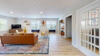 Located at 1608 Johnson Rd in Petersburg, VA, this single-floor space offers a thoughtfully designed interior that blends modern aesthetics with warm, welcoming touches. The expansive main living area is bathed in natural light streaming through multiple windows fitted with white plantation shutters, highlighting the light-toned hardwood flooring that runs throughout. The space is styled with mid-century modern furnishings, including a cognac leather sofa, navy velvet bench, and natural wood slab coffee tables anchored by a vibrant, multi-colored area rug. Recessed lighting complements the natural light, keeping the interior bright and inviting at all times. A dedicated seating and lounge zone flows seamlessly into additional areas, with open doorways and French doors connecting rooms for a cohesive, airy feel. Decorative open shelving, curated wall art, and tasteful accent pieces add character throughout. The kitchenette area features white cabinetry, light countertops, a stainless steel refrigerator, and a water cooler station, making it fully functional for daily use. A dining area with dark chairs sits adjacent to the kitchenette, completing the practical layout. This space presents a clean, contemporary atmosphere well-suited for a variety of uses.
