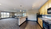Located at 6232 65th Ave N in Brooklyn Park, Minnesota, this single-floor community space offers a well-appointed and versatile layout designed for group gatherings and shared use. The space is divided into two distinct but connected zones: a fully equipped kitchen and an open lounge-style gathering area.

The kitchen features light maple cabinetry, dark granite-style countertops, a tile backsplash with decorative mosaic accents, and black appliances including a refrigerator, electric range, and over-the-range microwave. A peninsula counter with bar-height seating and a bold teal accent panel separates the kitchen from the main gathering area, with pendant lighting overhead adding a contemporary touch. Track lighting illuminates the kitchen zone effectively.

The gathering area is carpeted with a patterned, multi-tone carpet and furnished with round tables and black metal chairs, accommodating multiple seating arrangements. Large windows along the perimeter walls flood the space with natural light and offer views of the surrounding tree line. A sliding glass door provides access to an outdoor balcony, enhancing the connection to the outdoors. Recessed lighting and a textured ceiling complete the interior. The space transitions seamlessly from hardwood-style flooring near the kitchen to carpeting in the lounge area, creating a defined yet open flow throughout.