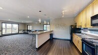 Located at 6232 65th Ave N in Brooklyn Park, Minnesota, this single-floor community space offers a well-appointed and versatile layout designed for group gatherings and shared use. The space is divided into two distinct but connected zones: a fully equipped kitchen and an open lounge-style gathering area.

The kitchen features light maple cabinetry, dark granite-style countertops, a tile backsplash with decorative mosaic accents, and black appliances including a refrigerator, electric range, and over-the-range microwave. A peninsula counter with bar-height seating and a bold teal accent panel separates the kitchen from the main gathering area, with pendant lighting overhead adding a contemporary touch. Track lighting illuminates the kitchen zone effectively.

The gathering area is carpeted with a patterned, multi-tone carpet and furnished with round tables and black metal chairs, accommodating multiple seating arrangements. Large windows along the perimeter walls flood the space with natural light and offer views of the surrounding tree line. A sliding glass door provides access to an outdoor balcony, enhancing the connection to the outdoors. Recessed lighting and a textured ceiling complete the interior. The space transitions seamlessly from hardwood-style flooring near the kitchen to carpeting in the lounge area, creating a defined yet open flow throughout.