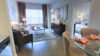 This single-floor residence showcases a thoughtfully designed open-concept layout that seamlessly connects the living and dining areas. The living room is anchored by rich hardwood-style flooring that extends throughout the space, lending warmth and a sense of continuity. Large windows dressed with floor-to-ceiling striped curtains allow generous natural light to fill the room, creating a bright and inviting atmosphere.

The living area is well-proportioned, comfortably accommodating a full sofa ensemble, upholstered ottoman seating, and a sleek rectangular coffee table with a chrome base. A bold, colorful abstract artwork serves as a focal point above a dark wood credenza, which provides additional storage and display space. Decorative table lamps throughout contribute to a layered lighting scheme that enhances the overall ambiance.

Adjacent to the living space, a glass-top dining table and a patterned accent chair add an elegant touch to the dining area. A glimpse into what appears to be a bedroom is visible through a doorway, suggesting a well-connected and functional floor plan. The overall aesthetic is modern and polished, with carefully curated furnishings and finishes that create a cohesive, contemporary feel.