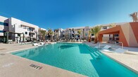 Located at 10340 S Decatur Blvd in Las Vegas, NV, this resort-style outdoor pool area offers a comprehensive recreational amenity designed for relaxation and socializing under the Nevada sun. The centerpiece is a generously sized swimming pool with crystal-clear turquoise water, complemented by a separate spa, both set against a backdrop of modern multi-story architecture with a clean, contemporary desert aesthetic.

The expansive sun deck is outfitted with numerous chaise lounges and umbrellas, providing ample space for sunbathing. Tall palm trees are strategically placed throughout, adding a tropical ambiance and natural shade. A dedicated lounge area features stylish outdoor sofas arranged around a glass fire pit table, ideal for evening gatherings.

An outdoor kitchen and grill station, sheltered beneath a modern pergola structure, provides convenient amenities for outdoor dining and entertaining. Additional seating areas with dining tables complete the social spaces. The pool area also includes a well-appointed pool house featuring accessible outdoor showers with stone tile accents and a dual water fountain station. The entire space is enclosed by sleek metal fencing and designed to maximize the open Las Vegas skyline views.