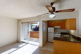 This single-floor unit offers a straightforward, functional layout with an open connection between the living area and kitchen. The living room is carpeted and bathed in natural light, largely courtesy of a full sliding glass door that opens directly onto a wooden deck with views of greenery beyond. A ceiling fan with a built-in light fixture is centrally mounted above the living area, complementing the textured ceiling throughout. Vertical blinds frame the sliding door, offering privacy and light control.

The kitchen sits adjacent to the living space and is well-equipped with warm-toned wood cabinetry offering ample upper and lower storage. Dark granite-style countertops provide a notable contrast, and white appliances — including a refrigerator, electric range, and dishwasher — complete the functional setup. A small window in the kitchen adds additional natural light.

The bedroom features carpeted flooring, neutral walls, and a sliding closet system that provides generous storage. A secondary closet is also visible nearby. The bathroom includes a vanity with matching dark granite countertop and wood cabinetry, a wall-mounted mirror, and a full tub-shower combination enclosed by sliding glass doors with tile surround. Tile flooring finishes the bathroom space cleanly.