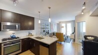 This single-floor space showcases a thoughtfully designed open-concept layout that seamlessly integrates the kitchen, dining, and living areas into one cohesive environment. The kitchen is appointed with rich dark cabinetry that contrasts elegantly with light quartz countertops, a classic white subway tile backsplash, and stainless steel appliances including a built-in dishwasher. A peninsula-style island with a double undermount sink provides additional workspace and casual seating, illuminated by two sleek pendant lights overhead. An open metal shelving unit serves as a stylish visual divider between the kitchen and living spaces, offering both display and storage opportunities. The living area is bright and airy, benefiting from large floor-to-ceiling windows and a glass door that floods the space with natural light. Light hardwood flooring runs throughout, enhancing the open and welcoming atmosphere. The overall design balances modern aesthetics with practical functionality, creating a comfortable and contemporary living environment well-suited for everyday use and entertaining.