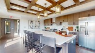 This thoughtfully designed single-floor residence seamlessly blends contemporary style with warm, natural materials throughout. The centerpiece of the home is a gourmet kitchen that commands immediate attention with its striking coffered ceiling detailed in rich wood tones and adorned with elegant amber glass pendant lights. Two-tone cabinetry in warm walnut and light wood finishes lines the walls, complemented by a dramatic marble-inspired tile backsplash and bright white quartz countertops. A large central island provides generous workspace and casual seating for several bar-height chairs, making it ideal for both everyday living and entertaining. Stainless steel appliances, including a professional-grade gas range beneath a sleek wall-mounted hood, complete the chef-ready setup. Wide-plank wood-look flooring flows throughout, grounding the open layout with warmth and continuity. Adjacent to the kitchen, a sun-drenched dining nook features large windows that frame scenic outdoor views, with direct access through sliding glass doors to a spacious outdoor deck. The deck offers ample room for multiple seating arrangements, including a dining set and lounge furniture shaded by a large umbrella, ideal for outdoor relaxation and al fresco dining. Together, these spaces create a refined, functional, and inviting home environment.